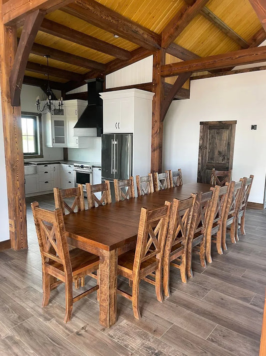 Large rustic wooden dining table with twelve matching wooden chairs in a farmhouse-style room with exposed beams and wood flooring